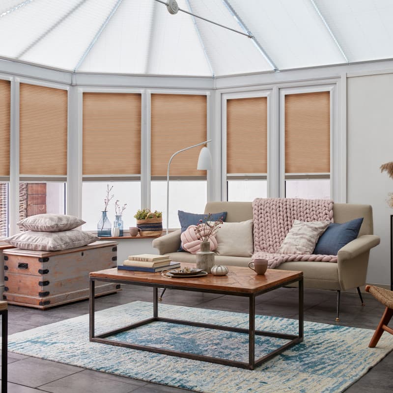 Beige pleated cellular blinds, lowered across tall conservatory windows, partially blocking and filtering daylight; in a bright sunroom living area with sofa, chunky pink throw, wooden coffee table.