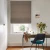 Taupe Roman blind, mostly lowered with soft folds, filtering daylight through a narrow gap; over a desk in a bright home office with vase of flowers, mirror and teal chair.