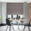 A taupe fabric Roman blind, fully lowered with horizontal folds, filtering daylight over glass French doors; dining area with white tulip table, four gray chairs, exposed brick and patio beyond.