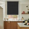 Dark charcoal woven roller blind fully lowered across window, showing horizontal textured weave and silver top rail; blocking light over a kitchen sink with brass tap, wood cabinets and marble countertop.