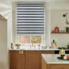 A grey-and-sheer zebra roller blind, lowered over the window with alternating opaque and translucent stripes, filtering light; kitchen with white walls, brass faucet, wooden cabinets and a brick courtyard view.