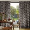Geometric-patterned pleated curtains, full-length and drawn partly aside revealing a central gap; framing a sunlit backyard with manicured lawn, trees, patio and a breakfast table set inside by the window.