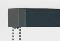 Dark gray metal roller-blind headrail and valance with two ball-chain pulls hanging; dark fabric blind tucked inside, mounted against a white wall in even studio lighting.