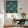 Floral Roman blind with teal background and yellow flowers, lowered over a sash window, partially filtering daylight; small home office desk beneath with mirror, vase of foliage, teal chair, walls.
