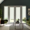 Double glass patio doors stand closed with white patterned roller blinds diffusing daylight, framing a living room with a glass coffee table, chevron rug, green armchairs, and potted plants.