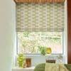 Roman fabric blind with olive-beige geometric wave pattern, partially lowered and filtering daylight; overlooks bedroom window with houseplants, glass vases, fan and bed with textured green bedding, view of shrubs.