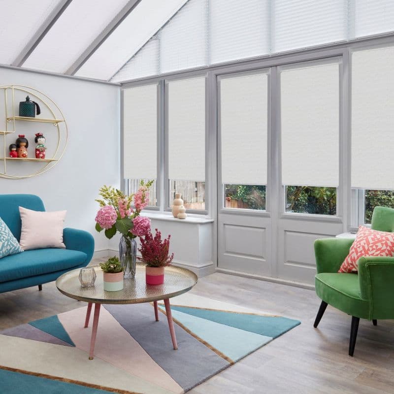 White roller blinds covering tall conservatory windows and triangular skylights, partially lowered to diffuse light, in a bright sunroom with teal sofa, green armchair, pastel rug and floral centerpiece.
