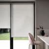 White semi-opaque roller blinds covering floor-to-ceiling black-framed glass doors, lowered to block view; soft daylight filters around edges into a modern kitchen nook with pink barstool and countertop.