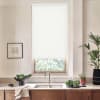 White roller blind covers upper portion of a window; a stainless faucet and sink sit below on a marble countertop with wooden cabinets, potted plants, greens, and framed art.