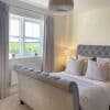 Floor-length pleated pale curtains hanging from a ceiling-mounted rod, pulled aside to frame a double-glazed window and admit daylight into a neutral, softly lit bedroom with a tufted bed.
