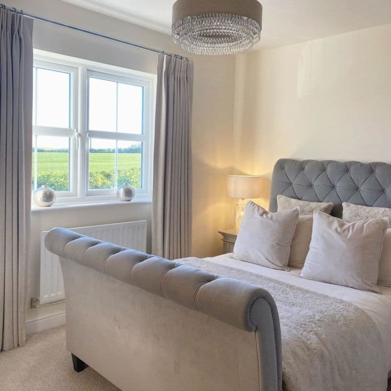 Floor-length pleated pale curtains hanging from a ceiling-mounted rod, pulled aside to frame a double-glazed window and admit daylight into a neutral, softly lit bedroom with a tufted bed.