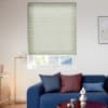 Light-green cellular blind, fully lowered over the upper window with horizontal pleats, filtering daylight; modern living room setting with blue sofa, cushions, floor lamp and partial outside roof view.