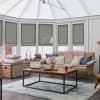 Pleated cellular blinds across tall bay windows, lowered to mid-height and filtering daylight; modern conservatory living room with vaulted glass roof, beige sofa, wooden coffee table and blue rug.