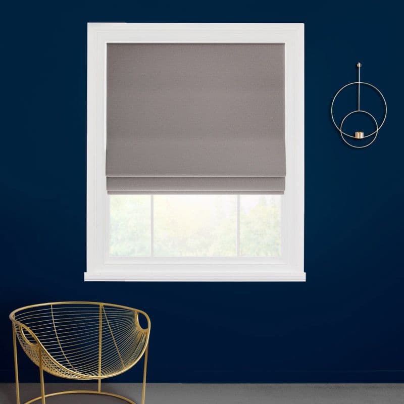 Grey fabric Roman shade lowered with visible folded tiers, filtering daylight; set in a white-framed window on a deep navy wall above a gold wire chair and circular wall sconce.
