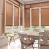 Brown horizontal Venetian blinds cover tall French-style windows and doors, lowered with slats mostly closed, filtering soft daylight into a bright dining nook with a wooden table, chairs, and garden view.