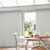 Light-gray vertical blinds, mostly closed with a narrow central gap filtering daylight; wooden dining table and wicker chairs in a bright conservatory-style kitchen, garden and shed visible outside.