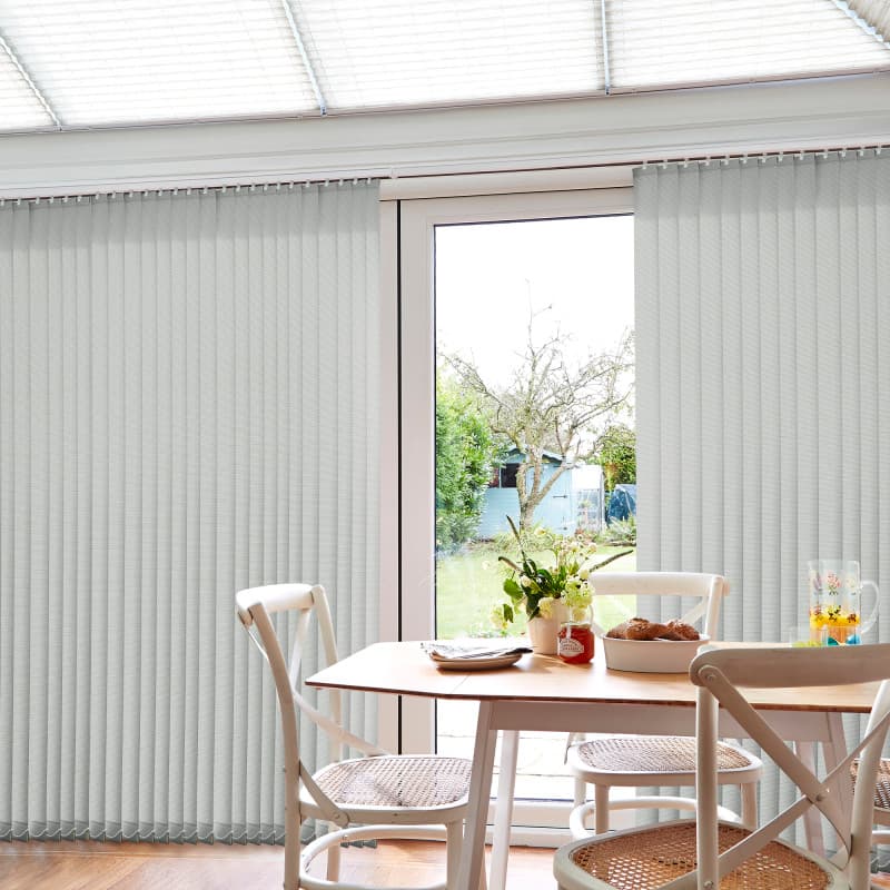 Light-gray vertical blinds, mostly closed with a narrow central gap filtering daylight; wooden dining table and wicker chairs in a bright conservatory-style kitchen, garden and shed visible outside.