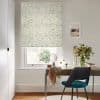 Roman blind of pale cream fabric printed with green-and-blue leaves, lowered over window and gently filtering daylight; small desk with teal chair, vase of foliage, framed seaside print.