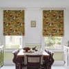 Two mustard-yellow floral Roman blinds, lowered over adjacent windows, patterned with red and blue flowers, filtering daylight into a dining room with wooden table, chairs and garden view.