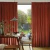 Heavy rust-red pleated curtains, pulled to each side and parted to reveal a glass door, filtering soft daylight over a dining table set for breakfast and a green backyard.
