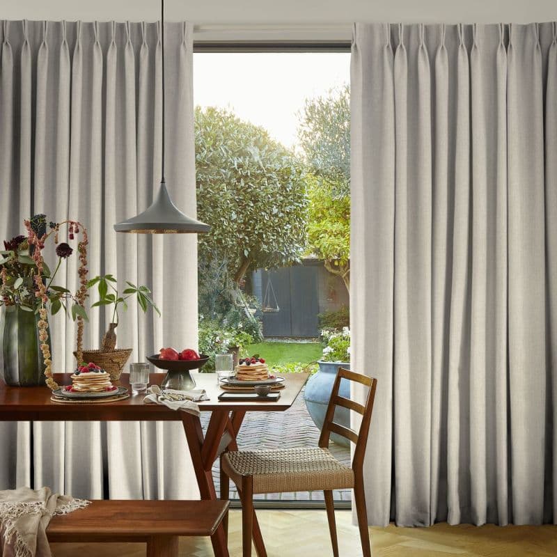 Beige floor-length pleated curtains, drawn partly aside to softly filter daylight through a sliding door; a wooden dining table with pancakes inside opens onto a sunlit garden path and shed.