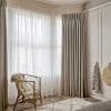 Floor-to-ceiling pleated beige curtains with sheer white voile panels across a corner bay window, filtering soft daylight into a neutral, cozy bedroom with rattan chair, wooden floors, rug and wardrobe.