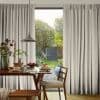 Full-length pleated beige curtains, slightly parted at center revealing glass door; framing daylight view. Wooden dining table set with plates, pendant lamp above; sunlit backyard garden with trees and lawn.