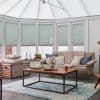 Honeycomb pleated blinds across tall conservatory windows, pale seafoam, lowered, leaving narrow bottom gap; filtering daylight. Context: bright sunroom with beige sofa, chunky pink throw, wooden coffee table and rug.