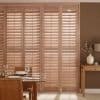Wooden plantation shutters cover floor-to-ceiling doors, closed with tilted slats filtering sunlight and casting striped shadows; warm dining area visible with glass table, place settings and side table with vase.