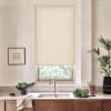 Beige roller blind covers most of a kitchen window above a stainless sink; surrounding countertop features wooden cabinets, a bowl of leafy greens, potted plants, and a framed wall sketch.