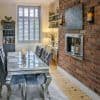 Object — white plantation shutters over a tall window with angled louvers; Action — mostly closed, filtering daylight; Context — dining room with mirrored table, tufted grey chairs, exposed brick wall and recessed fireplace.