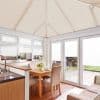 White pleated blinds cover roof panels, upper windows, and patio doors, closed and softly diffusing daylight into a bright conservatory-style dining area with wooden table, beige chairs, and garden view.