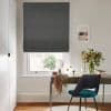 Dark grey textured Roman blind lowered, mostly closed and filtering daylight through a slim bottom gap; over a sash window above a desk with teal chair, vase of flowers.
