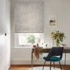 Light-grey floral-patterned Roman blind, lowered over the window, partially filtering bright daylight; a wooden desk and teal chair beneath, vase of flowers, neutral walls and framed beach print.