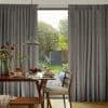 Floor-length gray pleated curtains, hung on a track and drawn wide to reveal a glass door, partially filtering light; breakfast table with pancakes and pendant lamp, green garden visible outside.