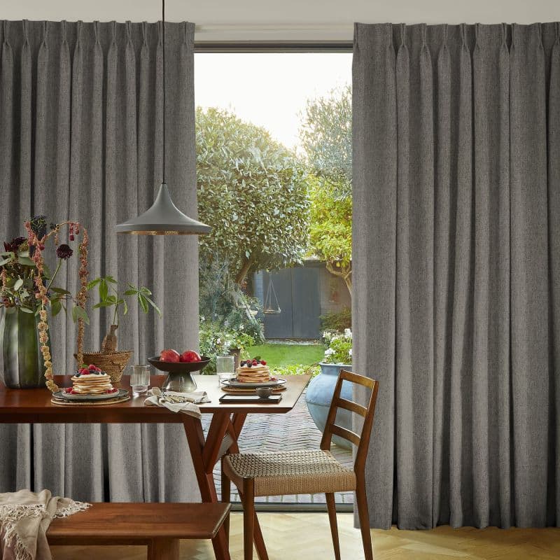 Floor-length gray pleated curtains, hung on a track and drawn wide to reveal a glass door, partially filtering light; breakfast table with pancakes and pendant lamp, green garden visible outside.