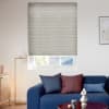 A light-gray pleated cellular blind, lowered with subtle horizontal ribs and a bottom gap, filtering daylight in a bright living room with blue sofa, cushions, floor lamp and coffee table.