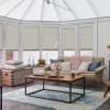 Light beige cellular pleated blinds, lowered across tall conservatory windows and slightly raised at lower edges, filtering soft daylight; cozy sitting room with sofa, chunky pink throw, wooden coffee table.
