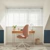 White horizontal Venetian blinds covering a wide window, slats mostly closed and filtering bright daylight; home office with wooden desk, pink swivel chair, lamp, plant, pale walls and blue alcoves.