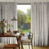 Full-length, pleated pale-grey curtains with leaf pattern, drawn open at center, framing a sunlit garden. Wooden dining table set for breakfast and pendant lamp in a cozy dining room.