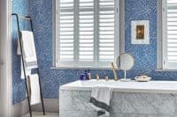 White plantation shutters covering the central window, wide horizontal louvers closed and softly diffusing daylight; bathroom interior with a marble-faced bathtub, brass fixtures, blue patterned wallpaper, and towel ladder.