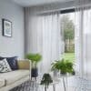 Sheer white, ceiling-mounted floor-to-ceiling curtains with pleats, partially drawn over sliding glass doors, filtering daylight into a living room with pale sofa, patterned tile floor, potted plants and lawn view.