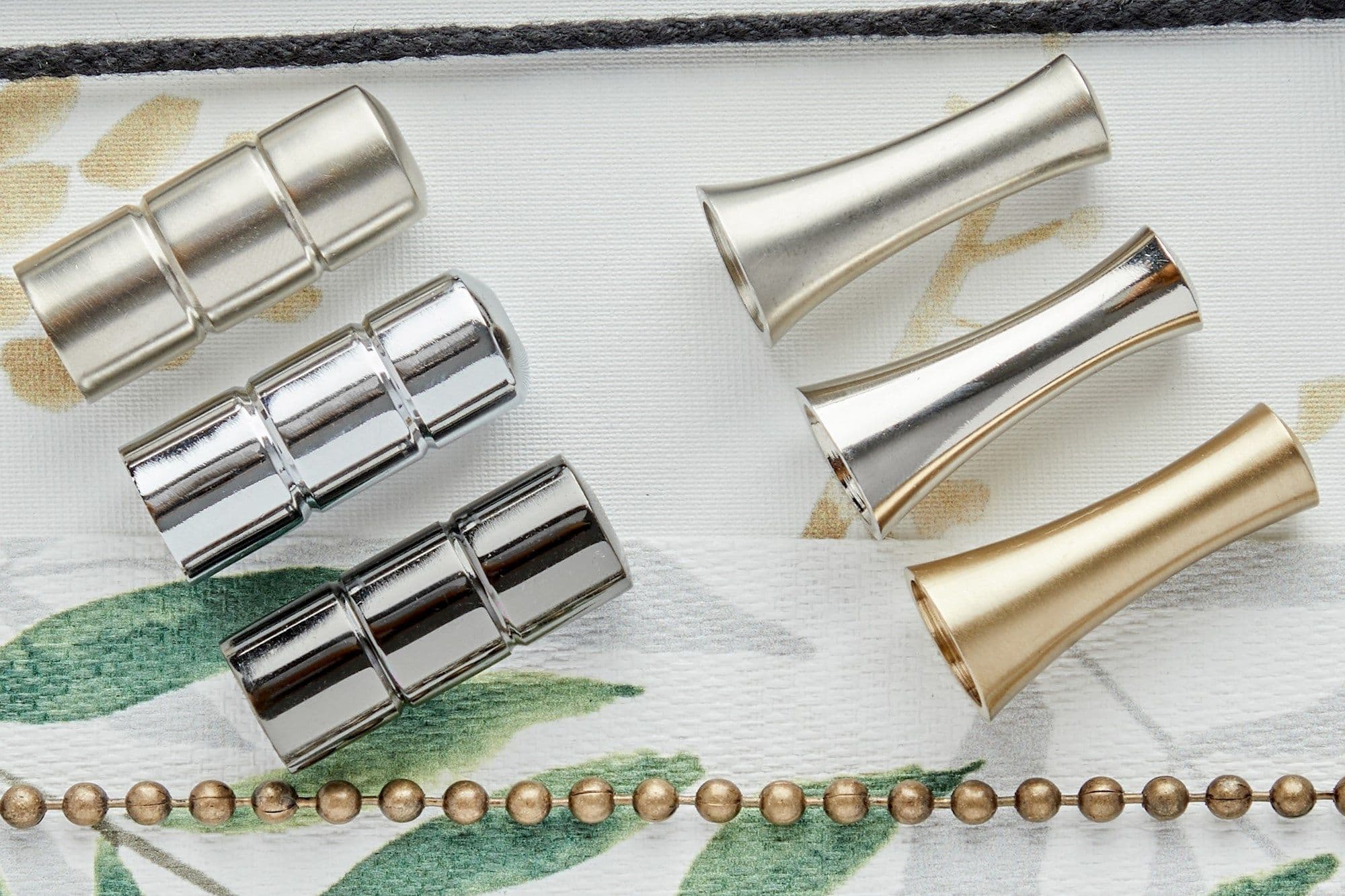 Metal curtain-rod components — three segmented chrome connectors (left) and three flared cone finials in silver and gold (right) — lie arranged on white leaf-print fabric under soft overhead light.