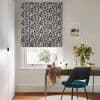 Black-and-white leaf-pattern Roman blind lowered over the window, filtering daylight; a wooden desk with teal chair, small round mirror and vase of foliage beneath, pale walls and hardwood floor.