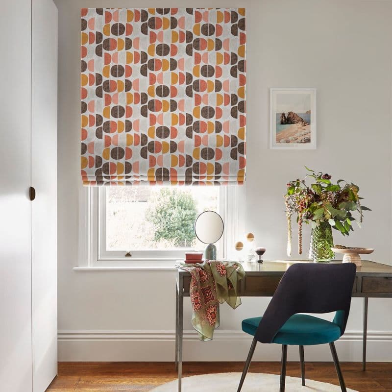 Roman blind with retro semi-circle pattern (mustard, pink, brown), partly lowered and softly filtering daylight; set over a home desk with chair, vase of flowers and framed seaside print.