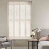 White plantation shutters covering a tall window, louvered panels closed and filtering soft daylight; set in a neutral-toned bedroom with upholstered bed, bedside table, lamp, and armchair.