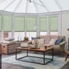 Pale green cellular blinds cover conservatory bay windows, lowered and filtering daylight; a neutral sofa with chunky pink throw and wooden coffee table sit on a blue rug.