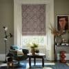 Patterned mauve Roman blind lowered over sash window, filtering daylight; cozy living room with green swivel armchair, dark carved coffee table, plants, floor lamp and framed portrait.