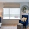 Beige fabric roman shade with a lowered white roller blind, both covering a large rectangular window; filtering soft daylight in a living-room corner with a blue armchair, ottoman, mirror.