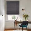 Dark textured Roman blind, fully lowered with a slight bottom gap, blocking light; mounted over a white sash window above a wooden desk with teal chair, mirror, vase of flowers, garden view.